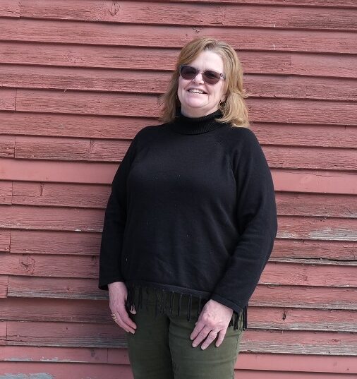 Tracy wears and dark sweater and sunglasses. She is smiling.