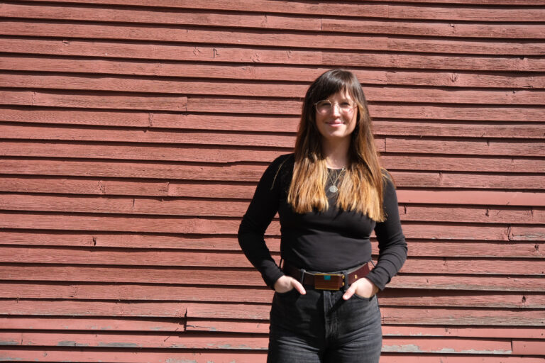 Molly has bangs and her hair has been balayaged from dark brown to light. She poses with her hands in her black jean front pockets.